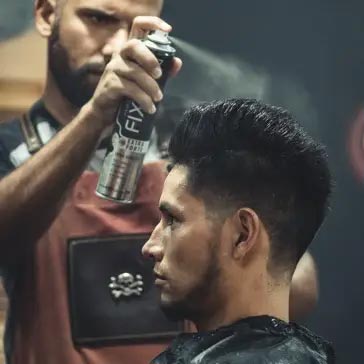 barber using styling spray on young man with black hair, short on sides and long in a quiff on top
